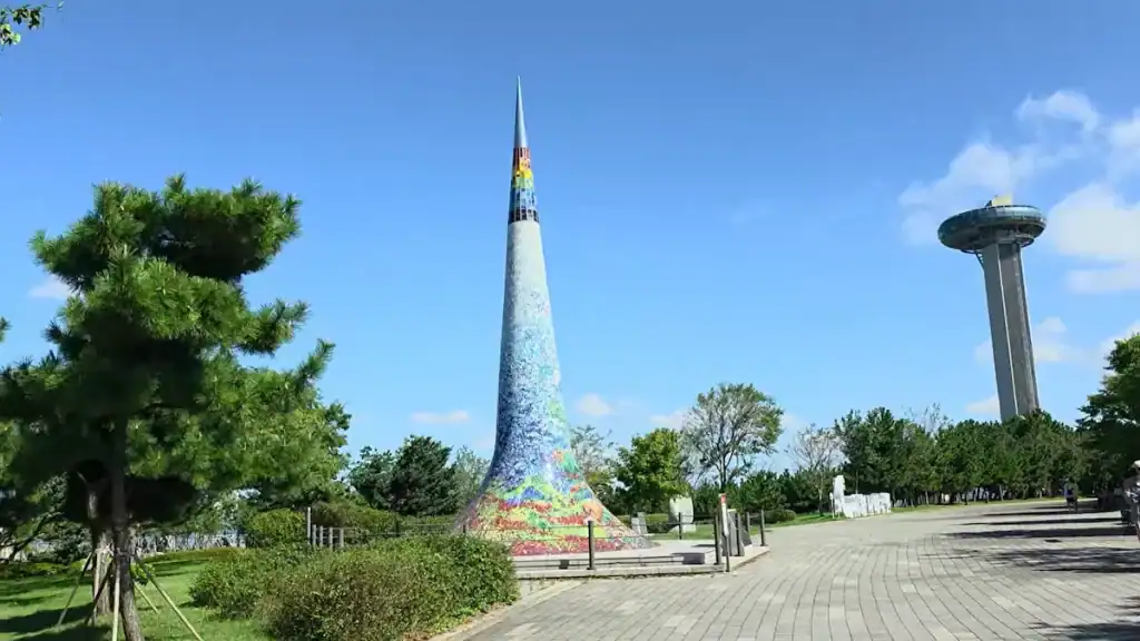 Colorful mosaic cone sculpture at Sihwa Tidal Power Plant park with observation tower in background along paved walkway and green trees