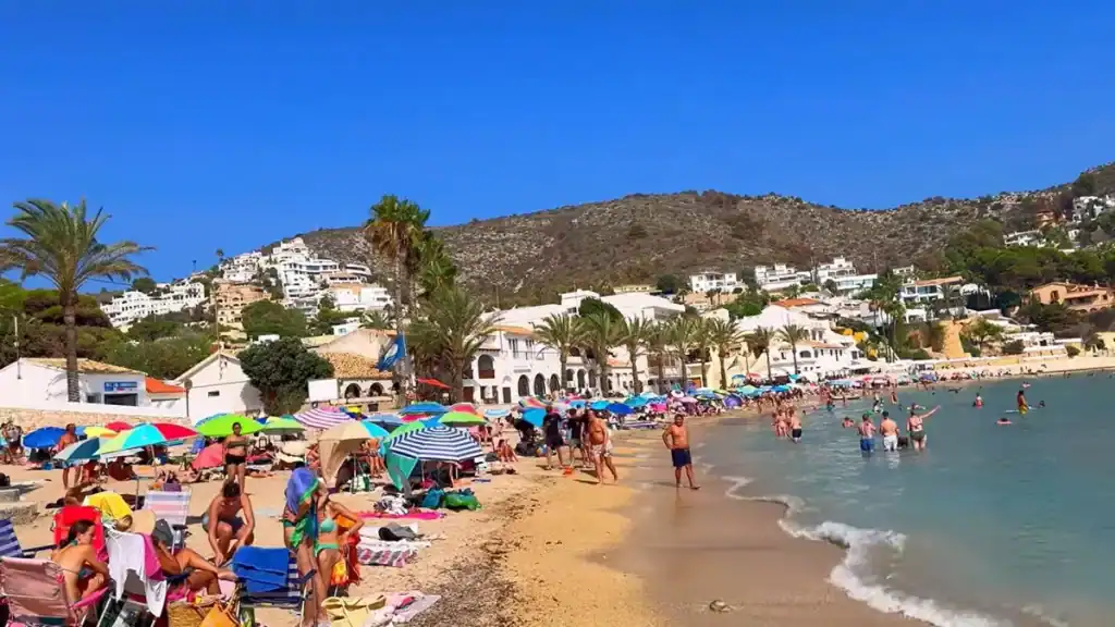 Crowded beach scene showing Moraira in August with colorful umbrellas and swimmers.