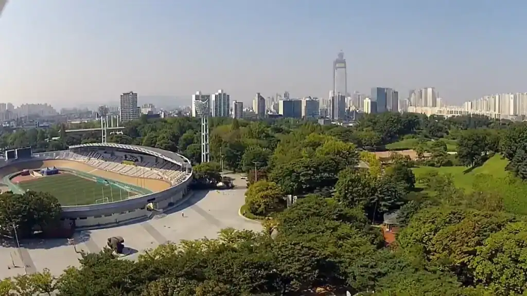 Aerial view of Seoul Olympic Park showing stadium, green spaces, walking paths, and city skyline with tall buildings