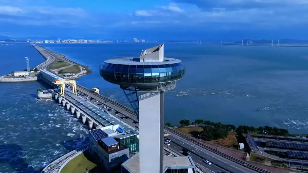 Sihwa Tidal Power Plant with distinctive circular observation tower, tidal barrage, roadway, and solar panels over blue water