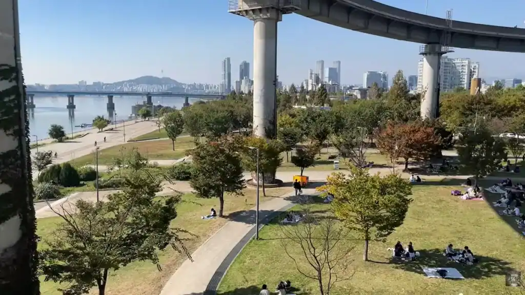 Ttukseom Hangang Park with green lawns, walking paths, people relaxing, Han River, bridges, and Seoul city skyline