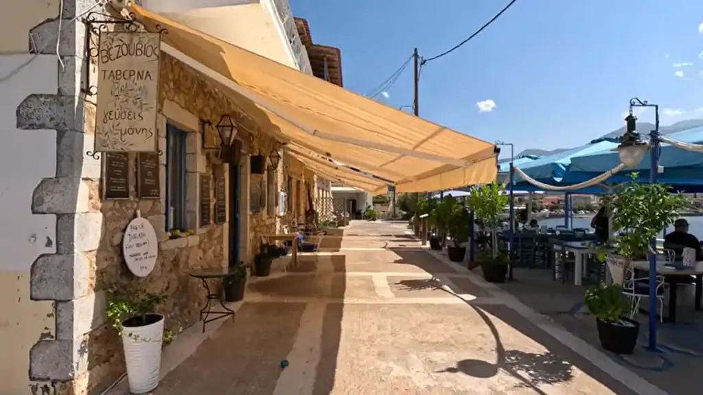 Stone-built taverna with a cream awning stretched over a harbourside terrace, Greek menu sign hanging from the corner wall and a few diners seated further along