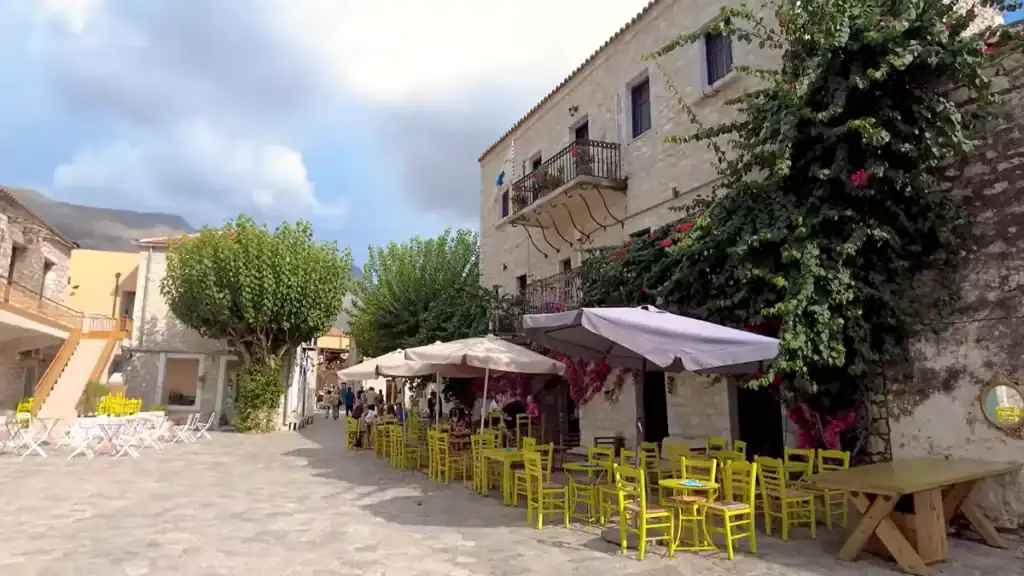 Yellow chairs and white parasols outside a stone-built cafรฉ on a sunlit village square, bougainvillea spilling across the faรงade above