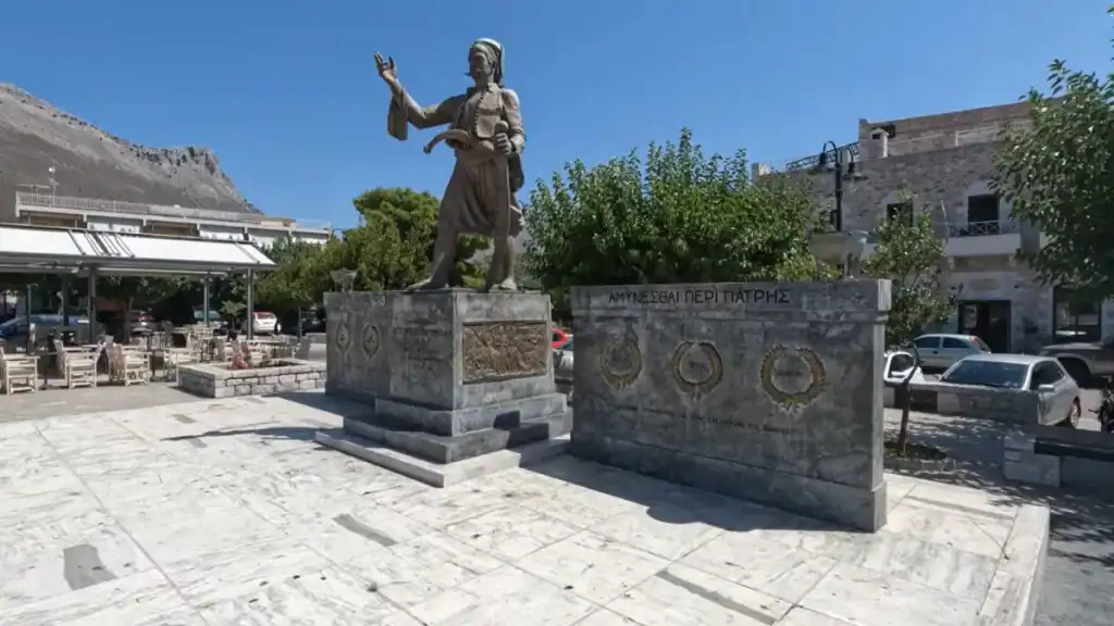 Bronze statue of a Maniะพั warrior raised on a stone plinth, one arm lifted, inscription reading ฮฮฮฅฮฮฮฃฮฮฮ ฮ ฮฮกฮ ฮ ฮฮคฮกฮฮฃ across the monument face, with a cafe terrace and limestone mountain behind
