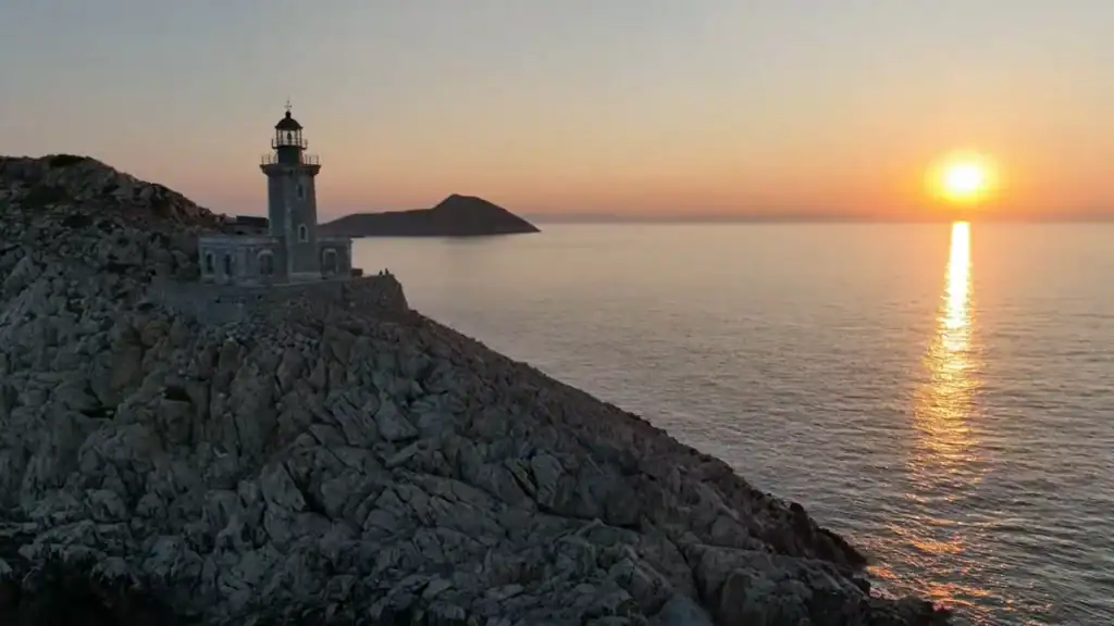 Lighthouse on a jagged rock promontory at sunset, the sun low over flat water with a small island silhouetted in the distance