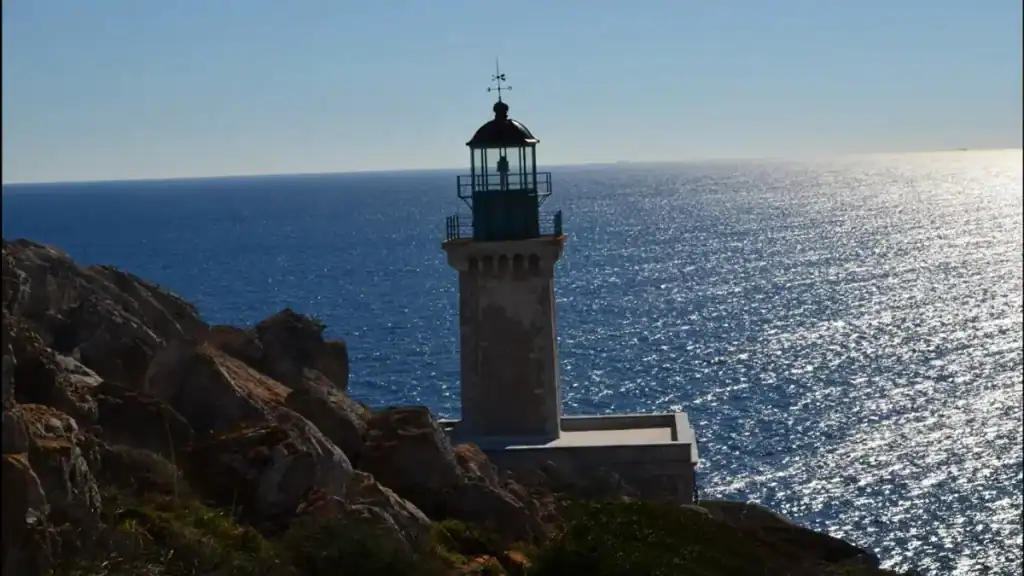 Weathered lighthouse tower rising from bare rock at Cape Tenaro, deep blue Mediterranean sea stretching to the horizon behind it