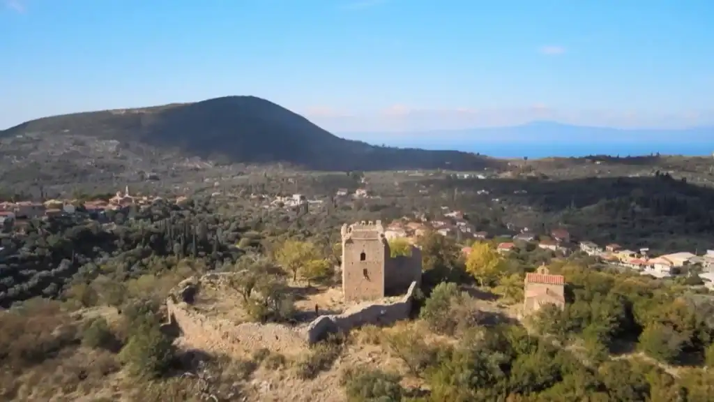 Ruined tower and enclosure walls of Zarnata Castle on a wooded hill, with a village and dark mountain behind it and the sea faintly visible on the horizon