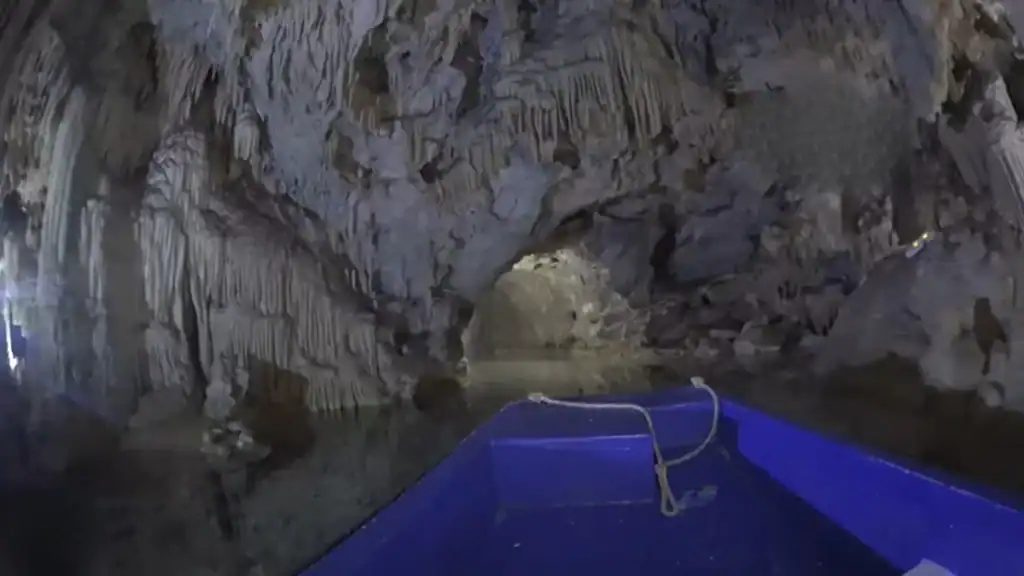 Blue boat moving through a flooded cave passage, stalactites hanging densely from the low arched ceiling above still water