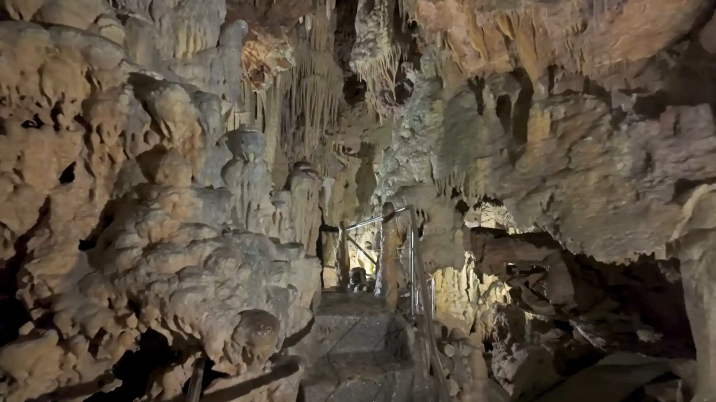 Stone steps descending between bulbous stalagmites and hanging formations on all sides, a metal handrail barely visible as the passage narrows toward a dim glow below
