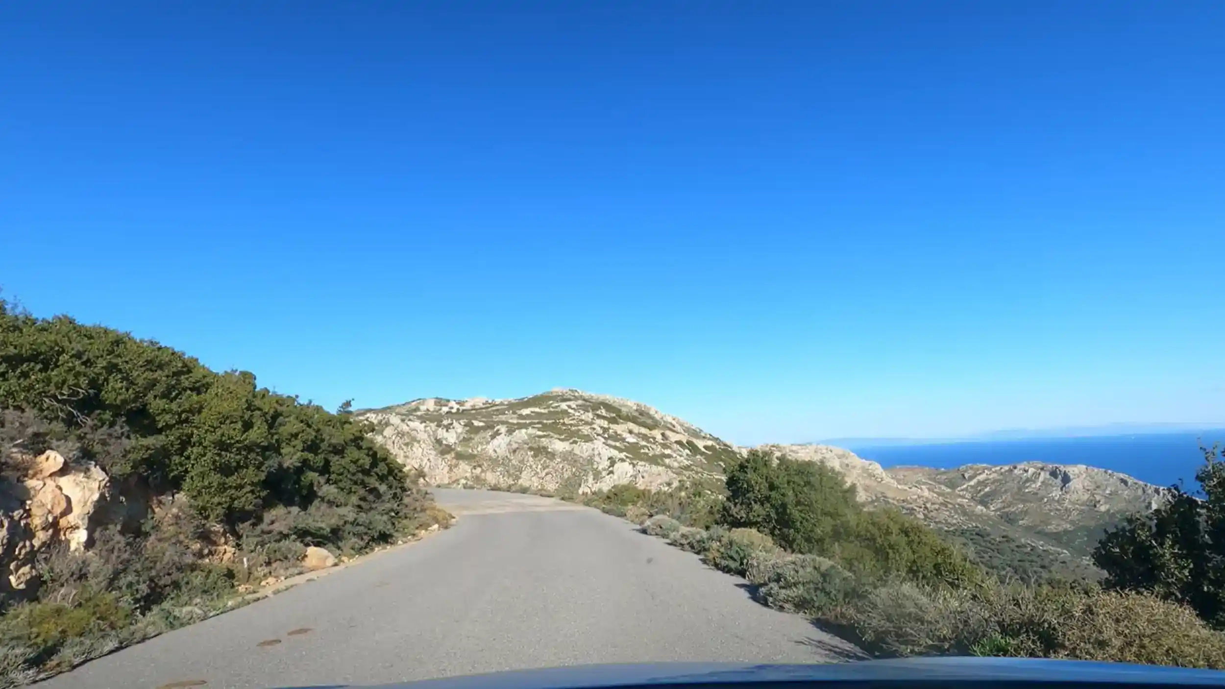 Narrow unmarked road climbing through rocky scrubland near Lagia, sea visible to the right between limestone ridges