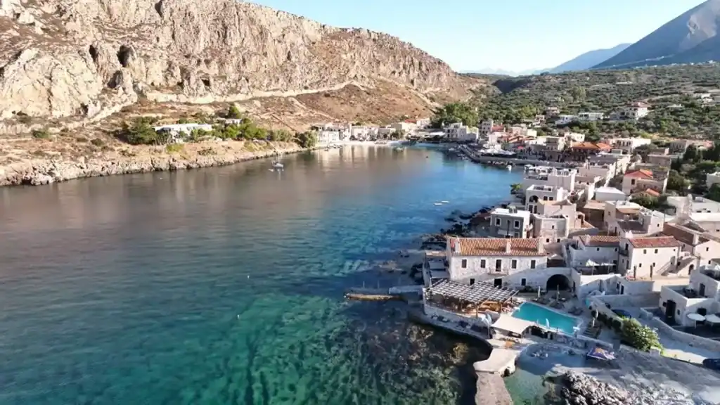 Stone-built village wrapping a sheltered inlet, turquoise water clear enough to show the rocky seabed, with a hotel pool cantilevered at the waterline and bare limestone cliffs closing off the bay to the left
