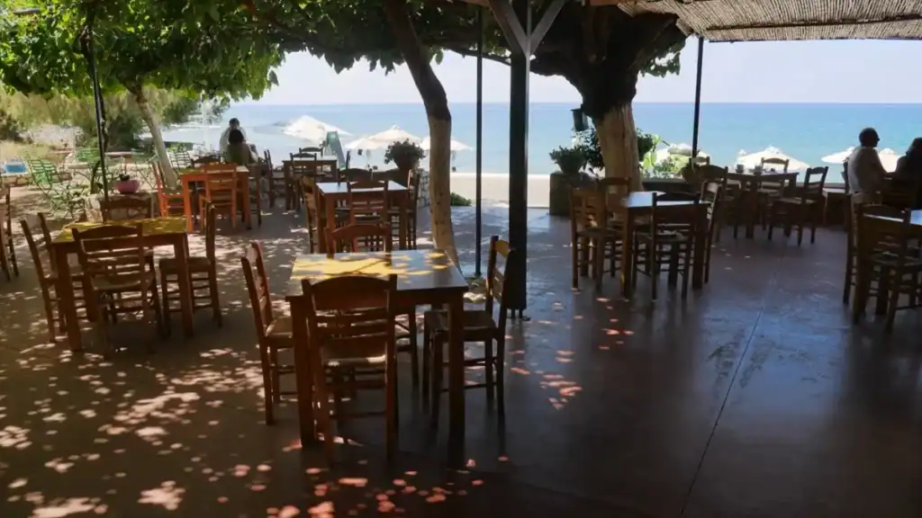 Open-air taverna mostly empty between service, wooden chairs and tables dappled with tree shadow, sea and beach umbrellas framed between trunks in the background