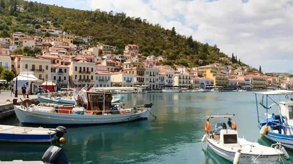 Wooden fishing boats moored in a calm harbour with a row of pastel-coloured neoclassical buildings along the waterfront and a densely wooded hill rising steeply behind the town