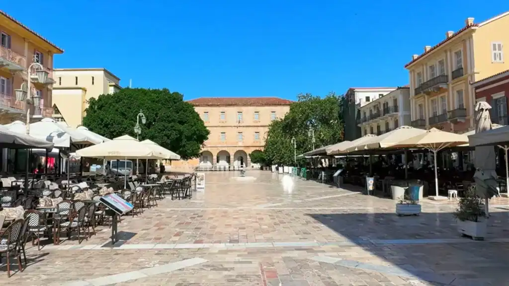 Wide marble-paved town square flanked by cafรฉ terraces under cream parasols, a neoclassical building with arched colonnades closing the far end