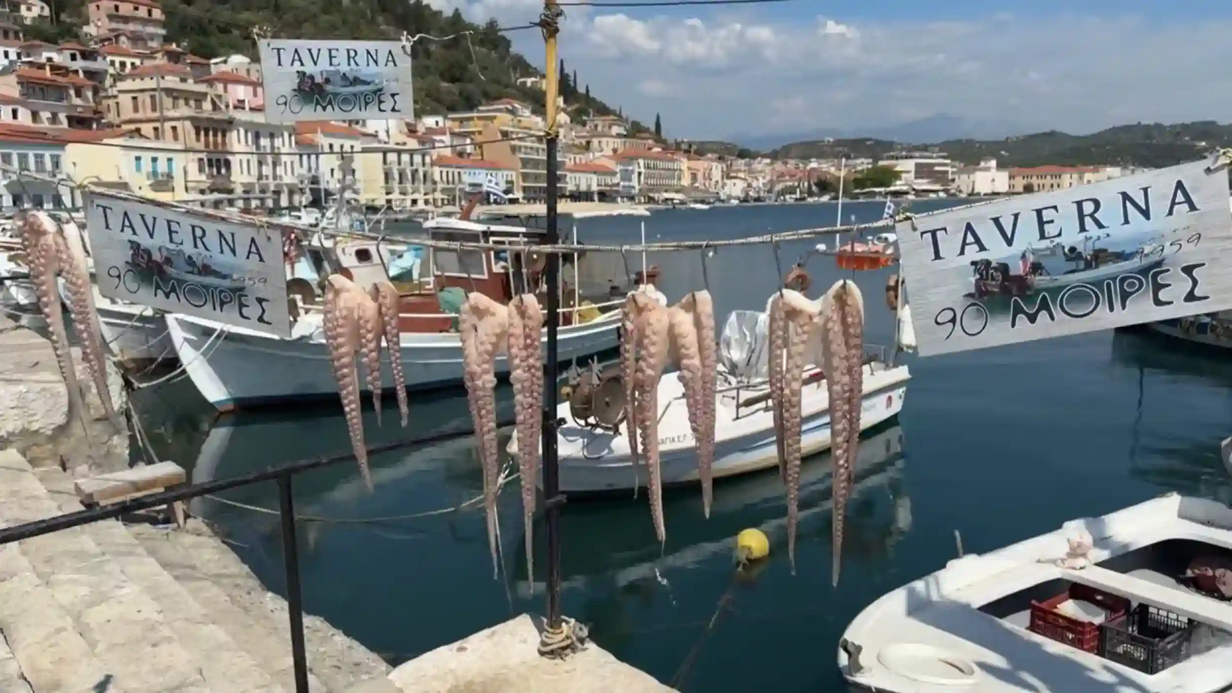 Octopus tentacles hung out to dry on a Mani Peninsula harbour line in front of moored fishing boats, taverna signs in Greek visible on either side, colourful waterfront buildings rising behind