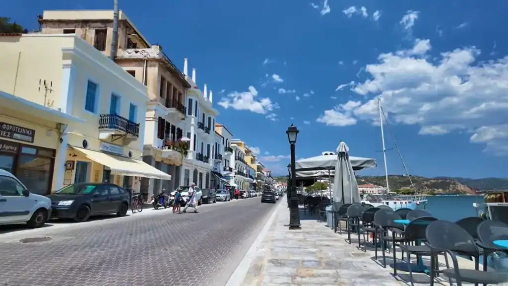 Cobbled waterfront promenade in Gytheio with neoclassical buildings lining the left side and empty cafe tables beside the harbour on the right