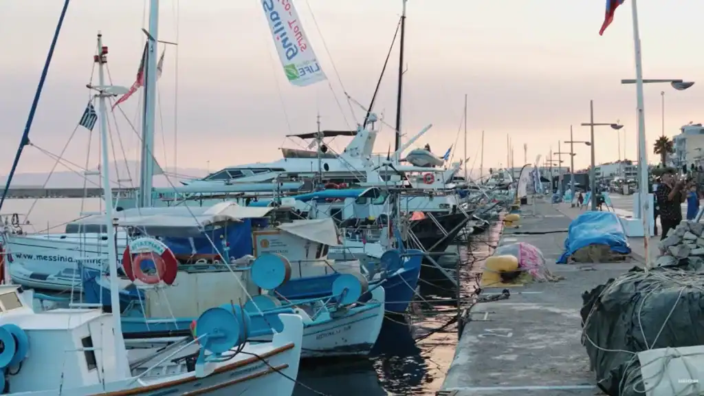 Fishing boats and sailing vessels moored along a harbourfront promenade at dusk, Greek flags visible among the masts, a banner for a sailing event hanging mid-frame