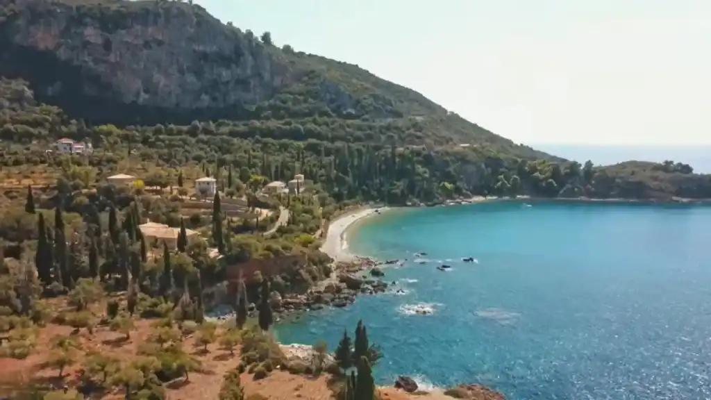 Curved pebble beach at Kalamitsi near Kardamyli, enclosed by turquoise water, with cypress trees, stone villas and a limestone cliff face rising steeply behind