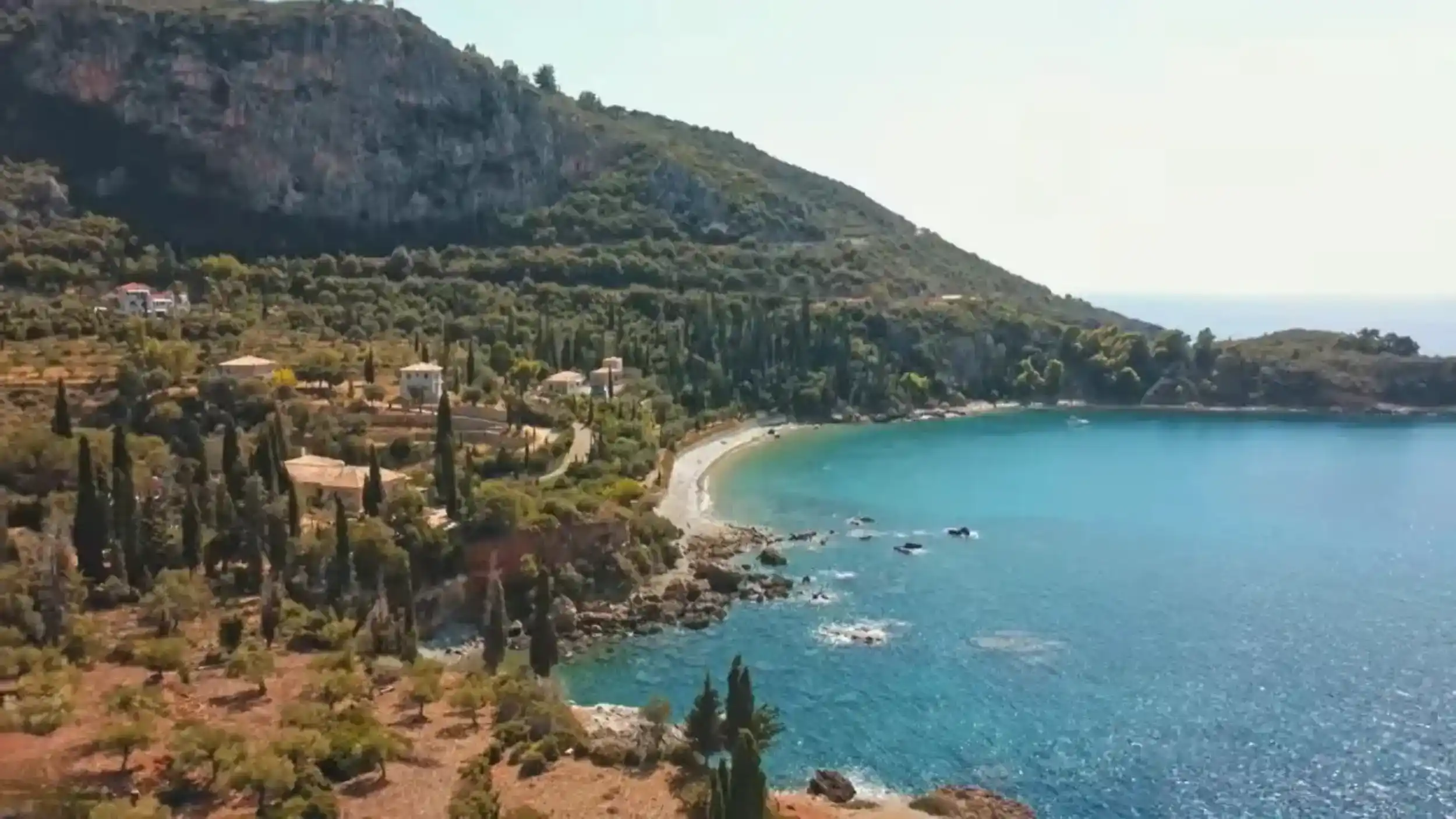 Curved pebble beach at Kalamitsi near Kardamyli, enclosed by turquoise water, with cypress trees, stone villas and a limestone cliff face rising steeply behind
