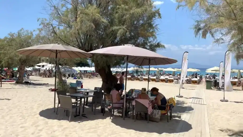 Packed sandy beach with rows of striped sunloungers and cream parasols, a lifeguard tower visible mid-frame and wooded hillside rising behind