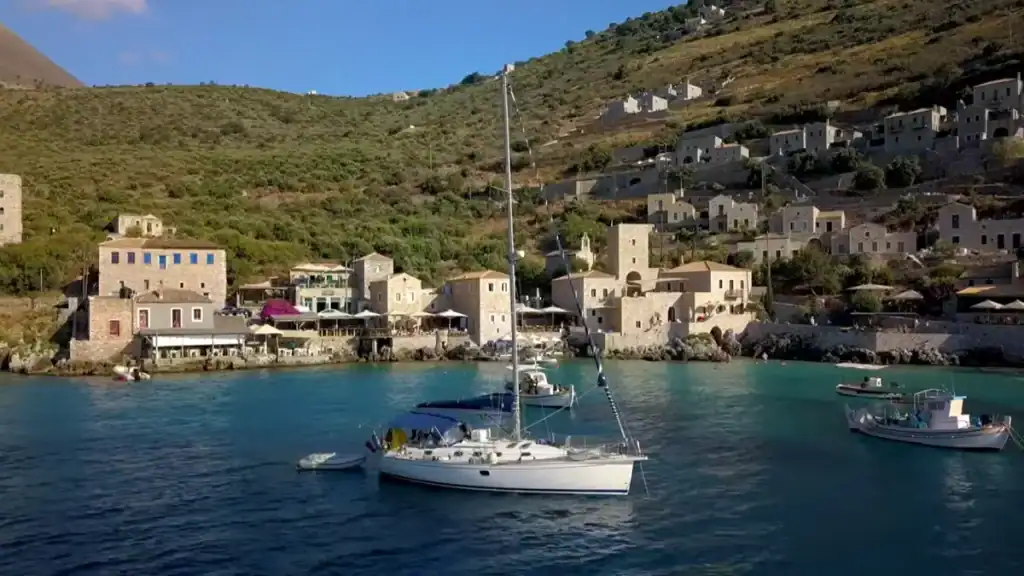 Sailboat anchored in a turquoise bay with stone-built village at the water's edge and a steep olive-covered hillside rising behind, smaller tower houses scattered across the slope