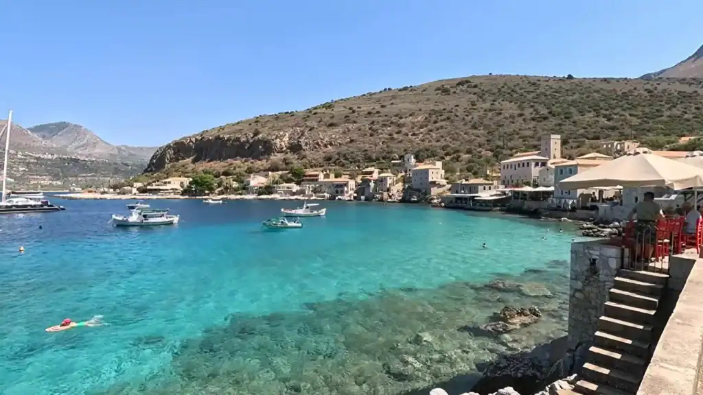 Intensely turquoise water filling a sheltered cove, a swimmer in the foreground, stone steps descending from a taverna terrace on the right, and tower houses clustered at the far shore beneath bare rocky hills