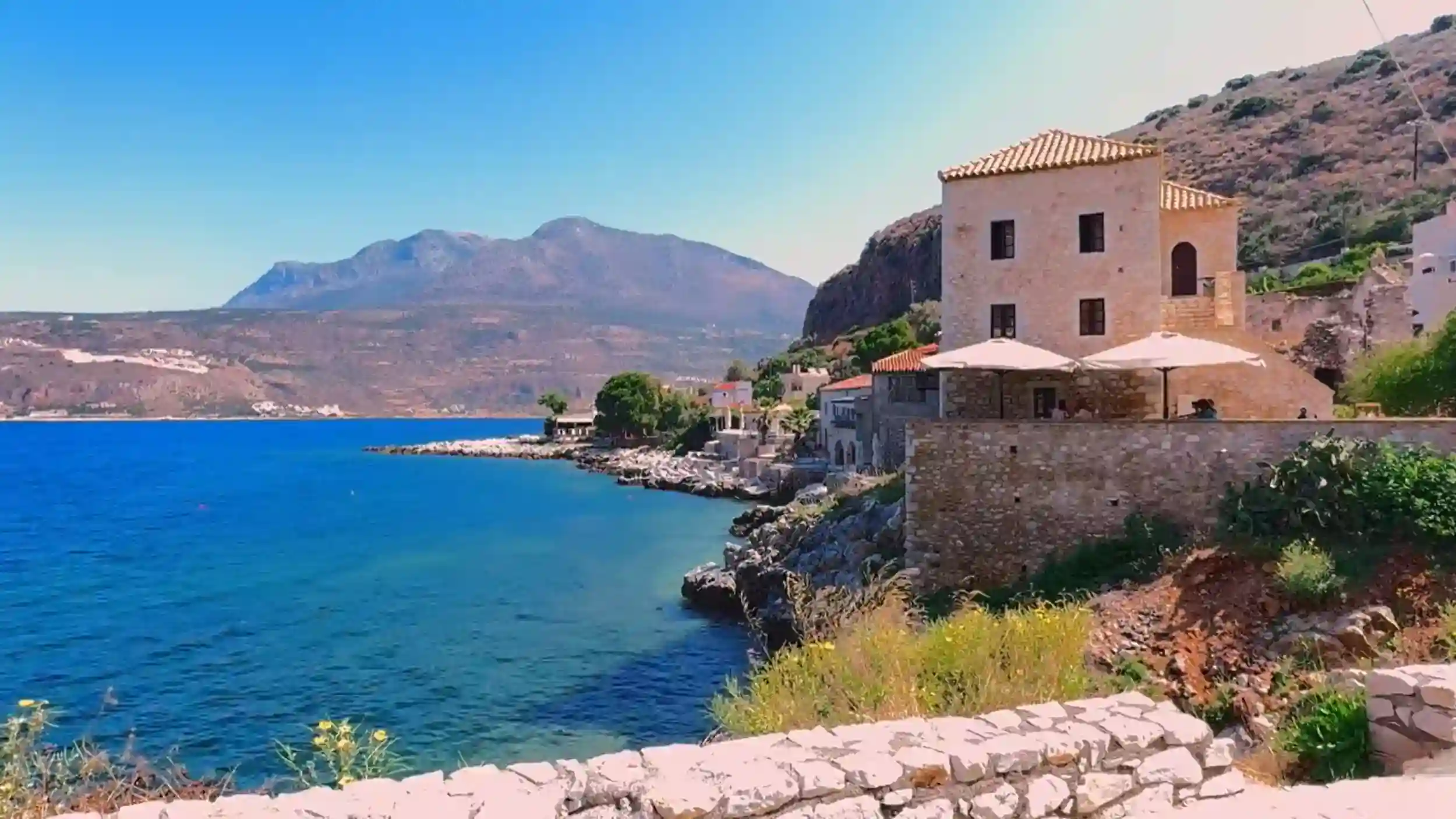 Sandstone tower house with a terrace and parasols sitting directly above rocky shoreline, deep blue gulf stretching to a broad mountain massif across the water