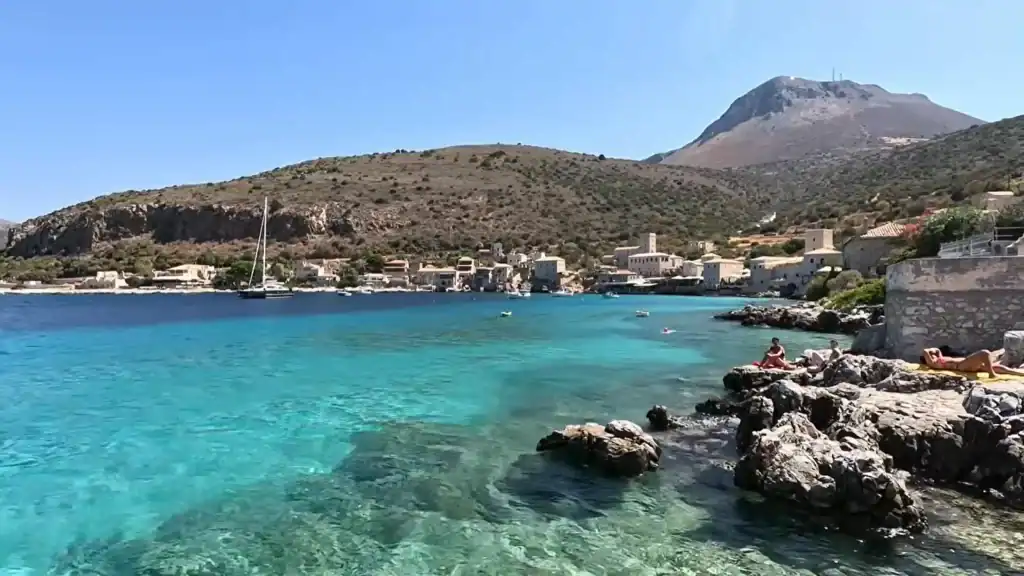 Turquoise water fills a sheltered bay at Limeni, with stone tower houses lining the far shore and sunbathers stretched across rocky foreground ledges