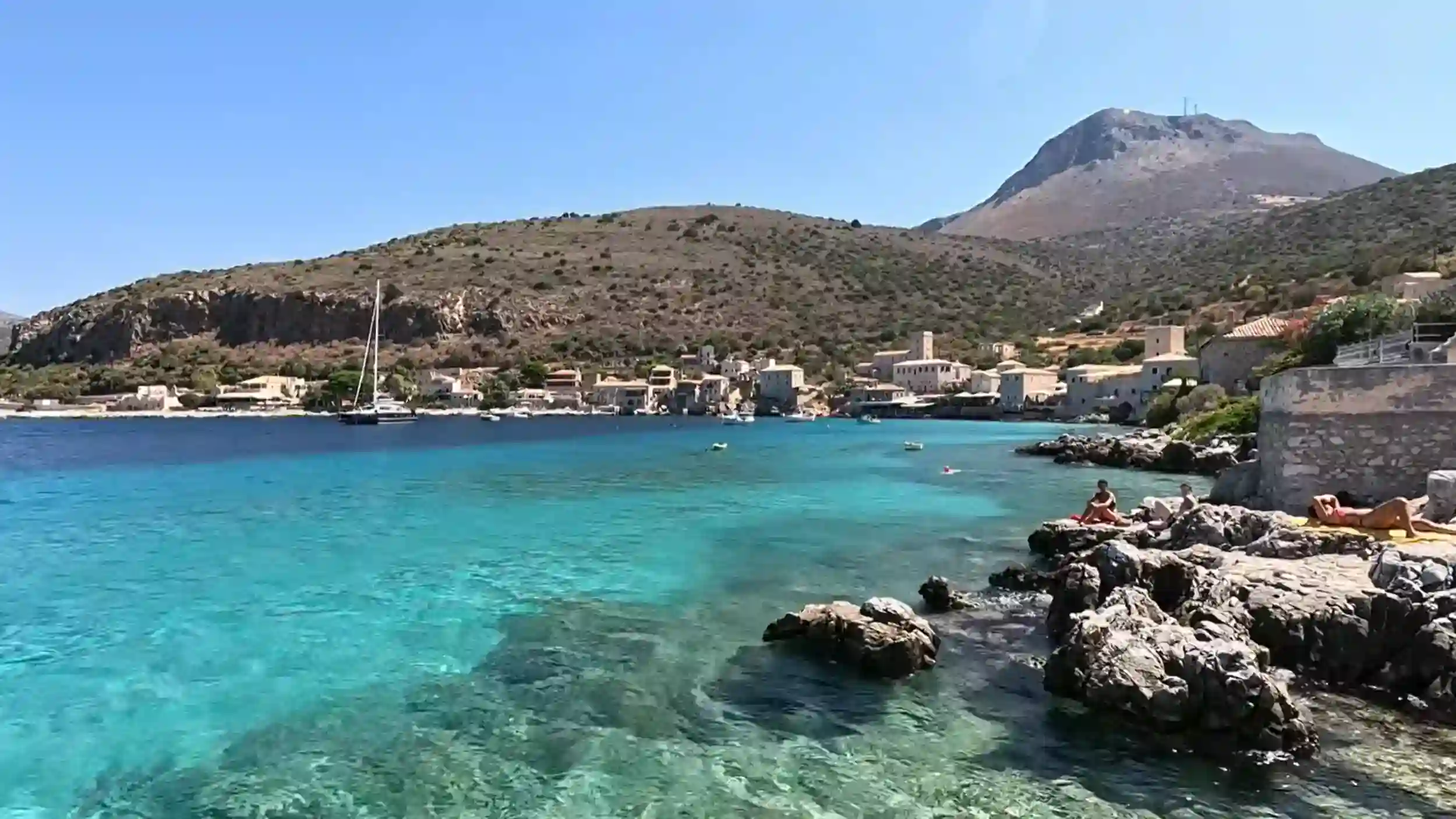 Turquoise water fills a sheltered bay at Limeni, with stone tower houses lining the far shore and sunbathers stretched across rocky foreground ledges