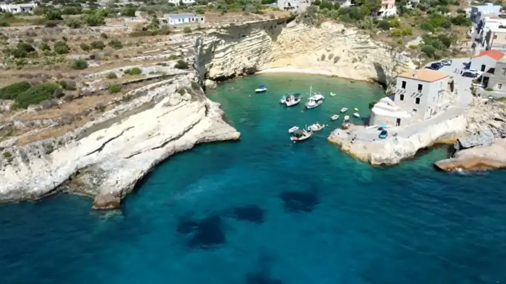 Small boats moored in a sheltered cove enclosed by pale layered limestone cliffs, stone buildings at the water's edge and a narrow sandy pocket at the far end