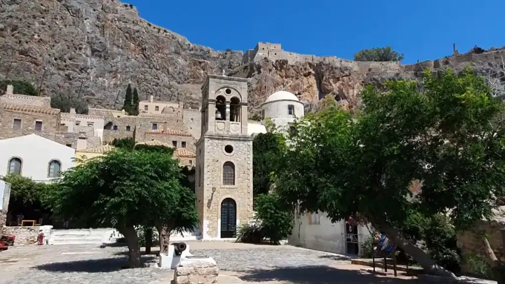 Stone bell tower rising above a shaded square in Monemvasia, with medieval buildings and a fortress wall climbing the cliff behind