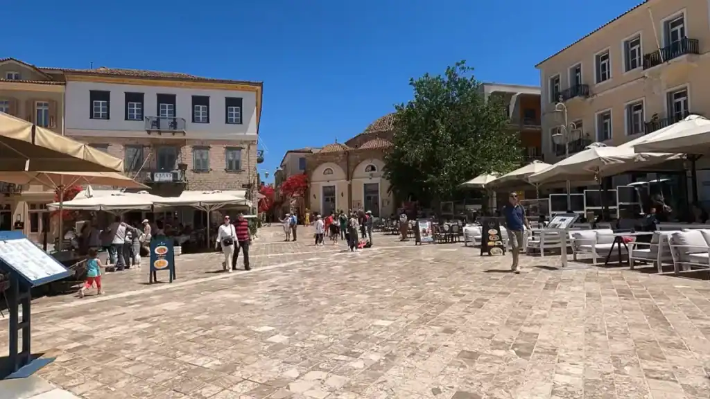Tourists cross a wide stone-paved square in Nafplio, with cafe umbrellas and a domed Ottoman-era building visible behind them