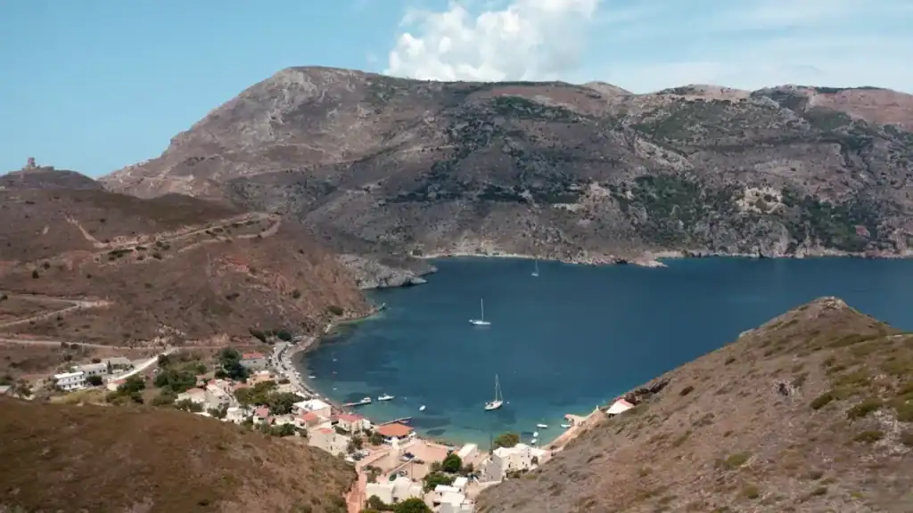 Enclosed bay with sailboats at anchor, a small whitewashed village curving along the waterfront and bare mountains rising steeply on all sides