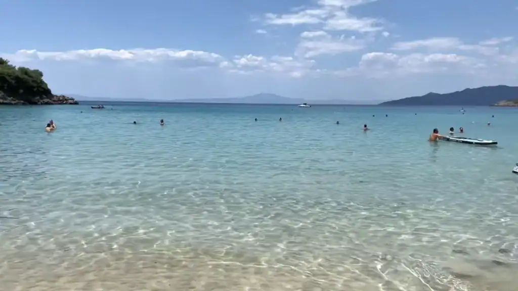 Shallow turquoise water lapping a sandy shore, swimmers scattered across a calm bay, distant headlands fading into haze under a partly cloudy sky