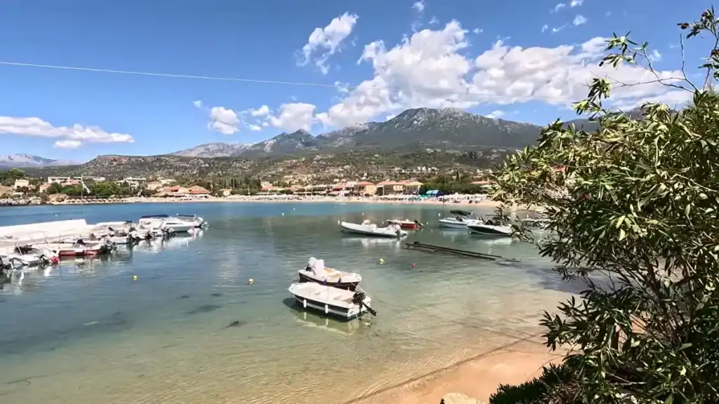 Shallow sandy bay with small motorboats anchored in clear water, a crowded beach with sunbeds visible across the cove, and a large mountain range dominating the horizon