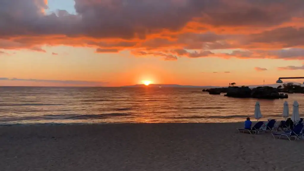 Deep orange sunset touching the horizon over a calm sea, heavy clouds lit from below, a lone figure on a beach lounger watching from the right