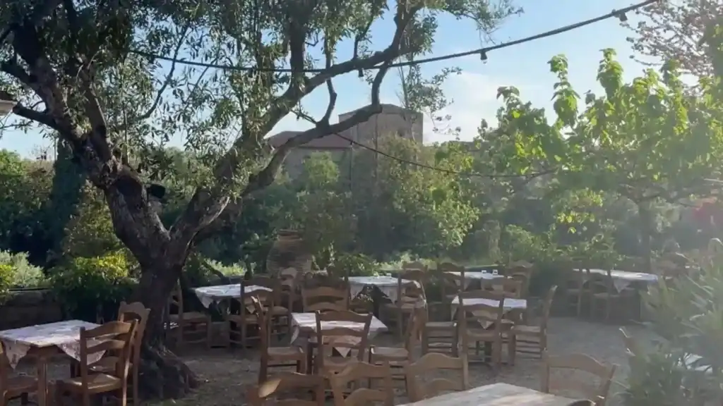 Empty wooden tables and chairs set beneath an olive tree in a garden taverna, with a stone tower visible through the foliage behind