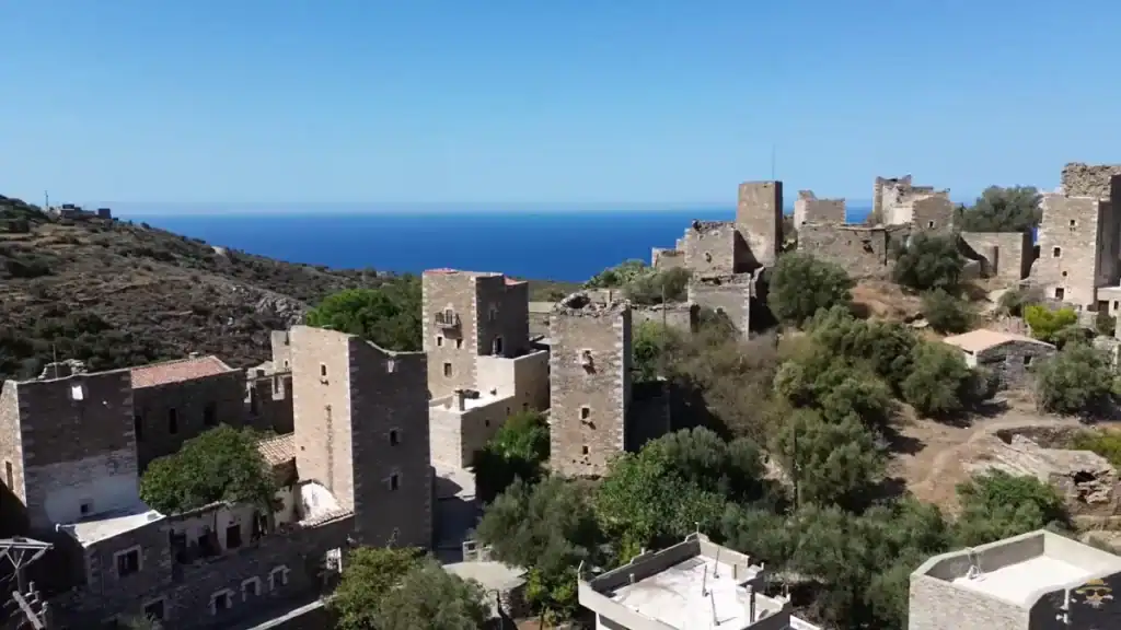 Dense cluster of tall stone tower houses in varying states of ruin rising above scrub-covered slopes, with deep blue open sea stretching to the horizon behind them