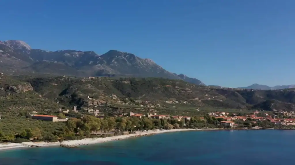 Aerial view of a curved pebble beach fronting a tree-lined village, the Taygetos range rising steeply behind with residual snow on the higher ridges