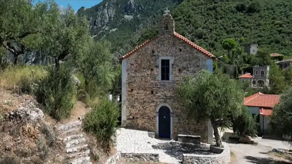 Small stone chapel with blue doors and a terracotta roof, set among olive trees with forested mountain slopes rising behind