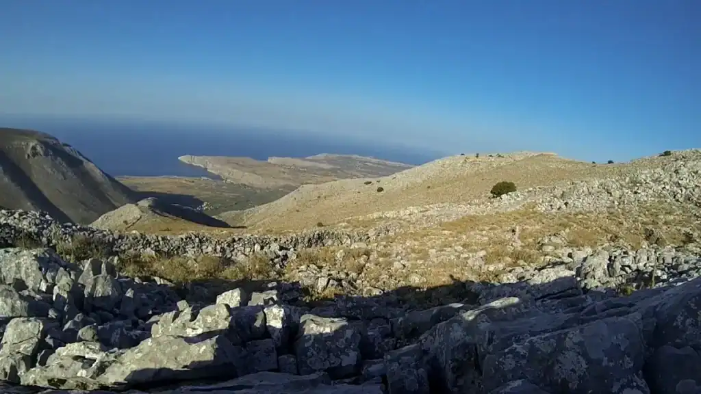 Rocky hillside path above a coastal headland, with a dry-stone wall cutting across sparse scrubland and the sea stretching flat to the horizon — hiking the Mani Peninsula in open, windless stillness