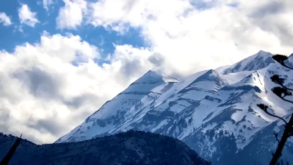 Snow-covered upper ridges of Mount Taygetos under a partly cloudy sky, with dark forested foothills along the base