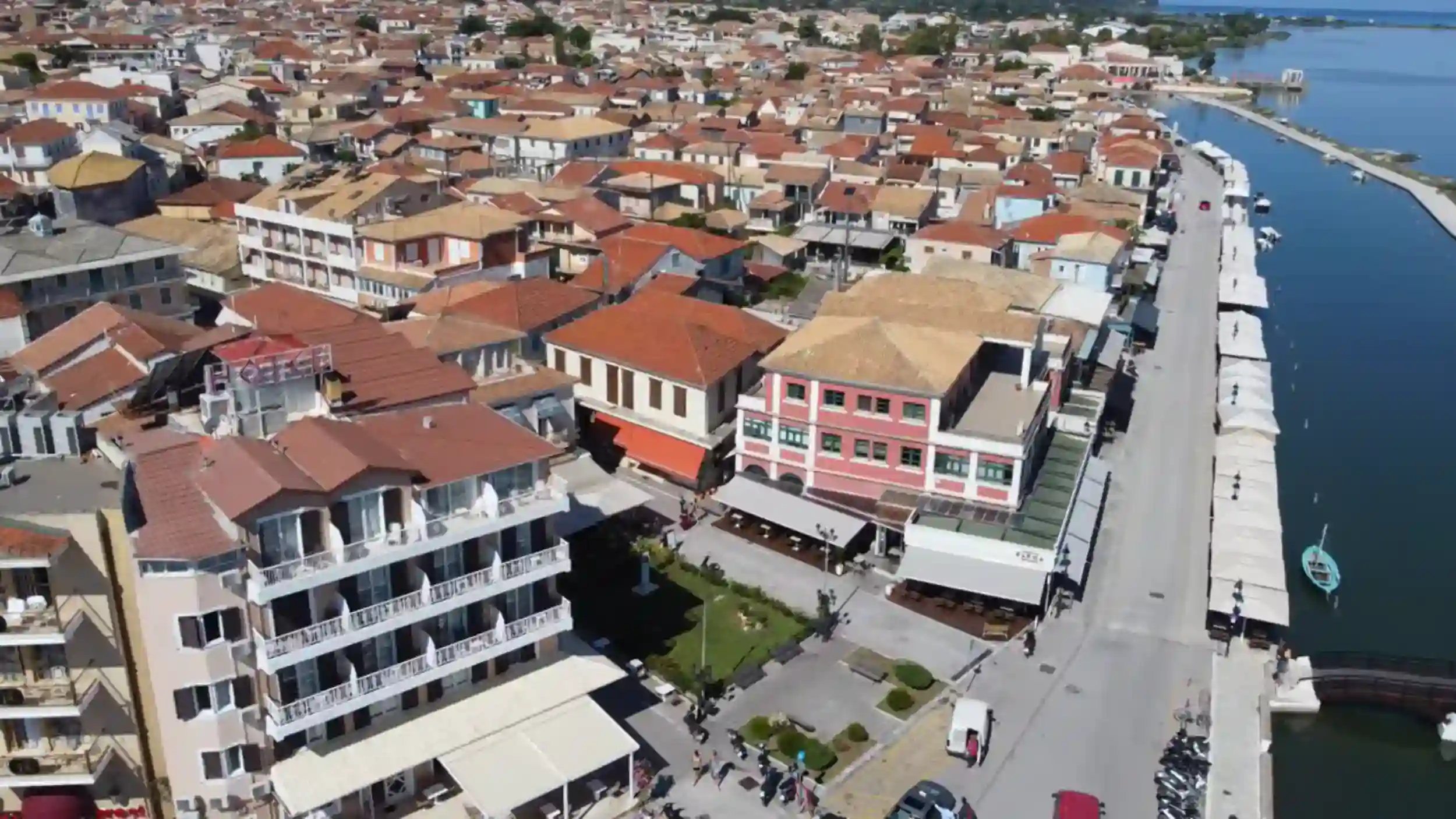 Densely packed terracotta rooftops of Lefkada town spread toward a calm waterfront promenade, with a small turquoise boat moored alongside the quay.