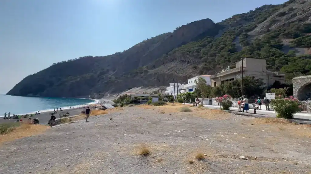 Small coastal village with white buildings beside a dark pebble beach, steep tree-scattered mountain dropping directly to the water's edge.