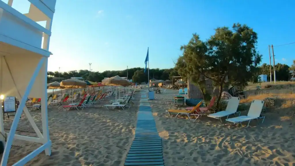 Agios Apostoli beach near Chania in December with colourful sun loungers and straw parasols lined up on sandy shore, a wooden boardwalk cutting through towards the trees