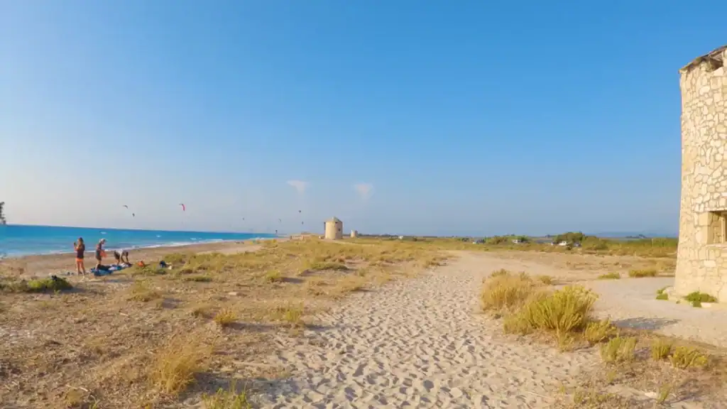 Old stone windmills standing on the flat sandy peninsula of Agios Ioannis Beach in Lefkada in September, with kitesurfers airborne over the water and a ruined mill wall in the foreground