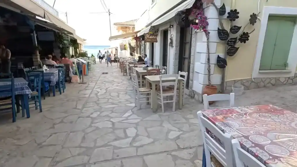 Stone-paved pedestrian lane with taverna tables set on both sides, the sea just visible at the far end where the street opens out, Maistros Taverna signed on the right