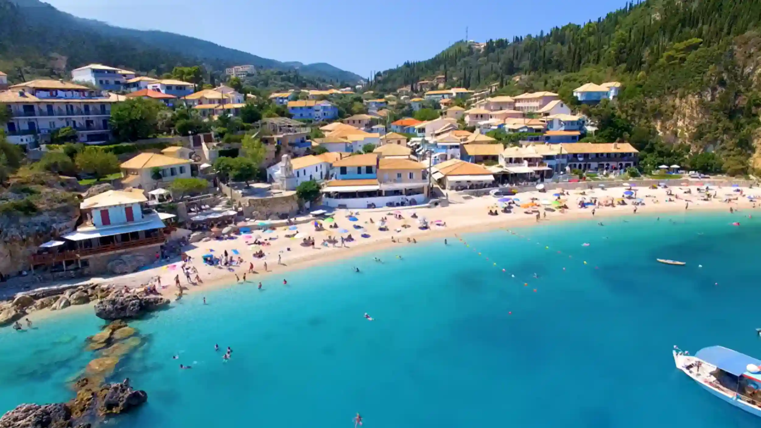 Agios Nikitas village and its sandy beach from above, terracotta-roofed buildings packed into a wooded hillside behind turquoise water busy with swimmers