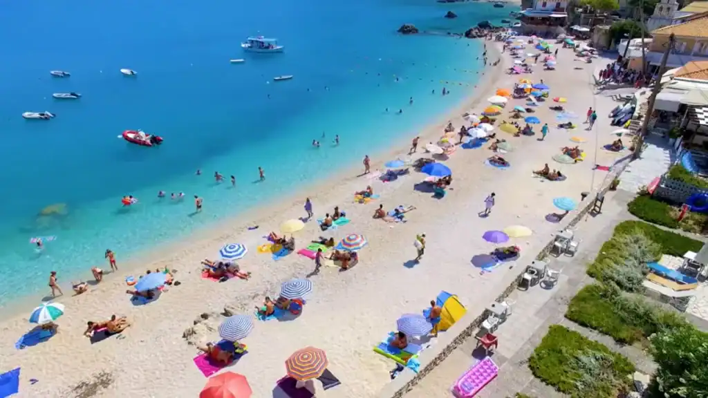Packed with colourful beach umbrellas and sunbathers, the Agios Nikitas beach curves alongside a promenade wall, boats moored in the shallow turquoise bay beyond