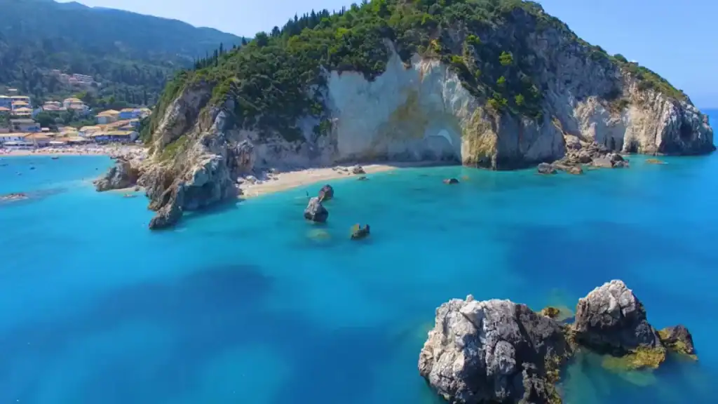 A densely wooded limestone headland drops to a secluded narrow beach, sea caves visible in the cliff face, scattered rock formations rising from vivid blue water in the foreground